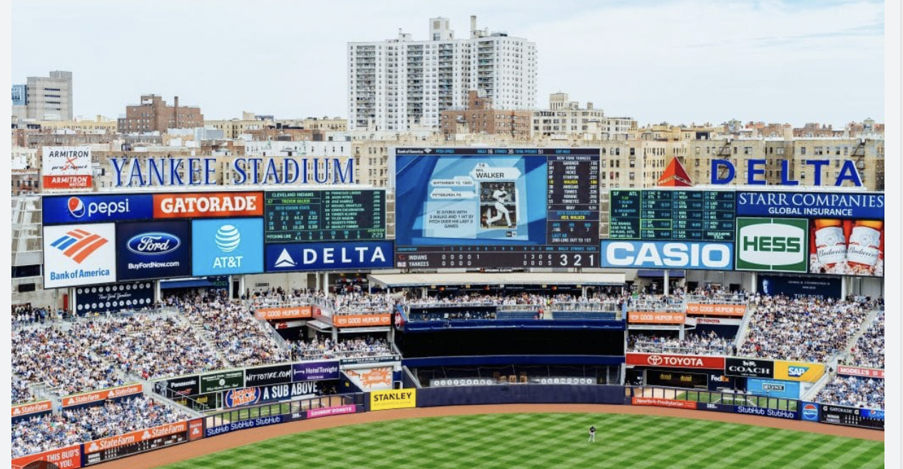 Crowd-filled Yankee Stadium during a baseball game with scoreboard and sponsor ads from brands like Pepsi, Delta, and Bank of America