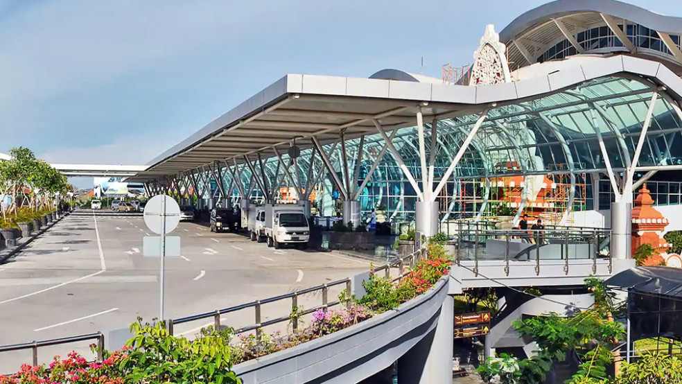 Modern exterior view of Bali Ngurah Rai International Airport with glass architecture, lush landscaping, and roadway access for passenger drop-off.