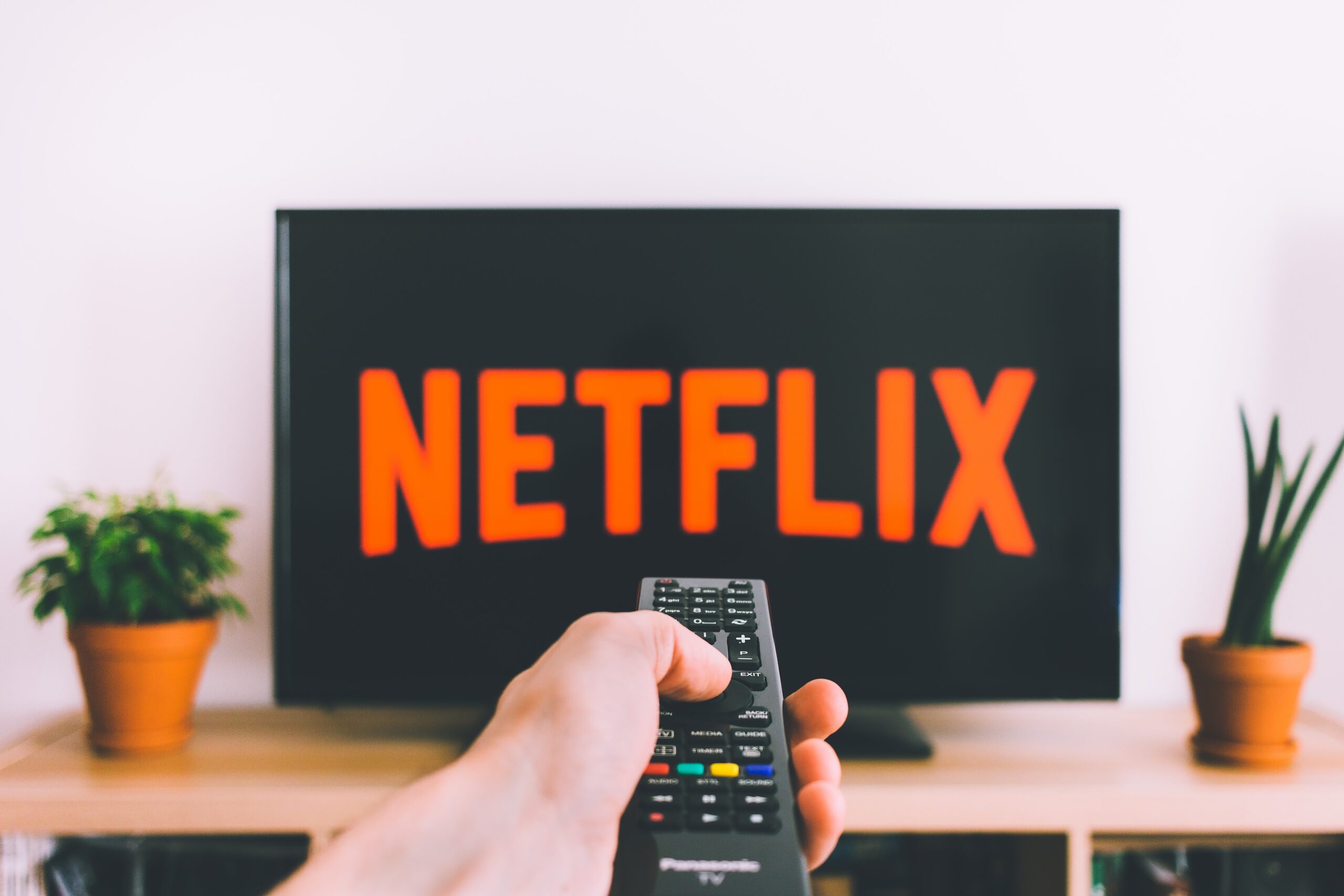 Hand holding remote control in front of TV screen displaying Netflix logo