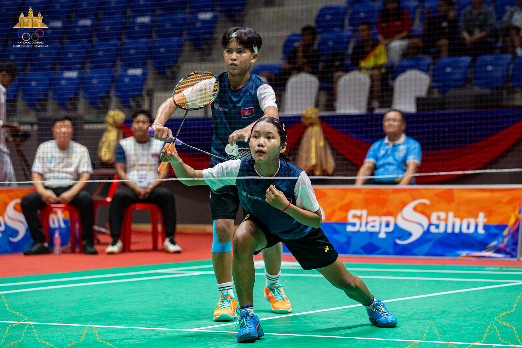 Cambodian mixed doubles badminton players in action during a competitive match, with the girl in front poised for a shot and the boy behind ready with a shuttlecock, at an indoor stadium with spectators.