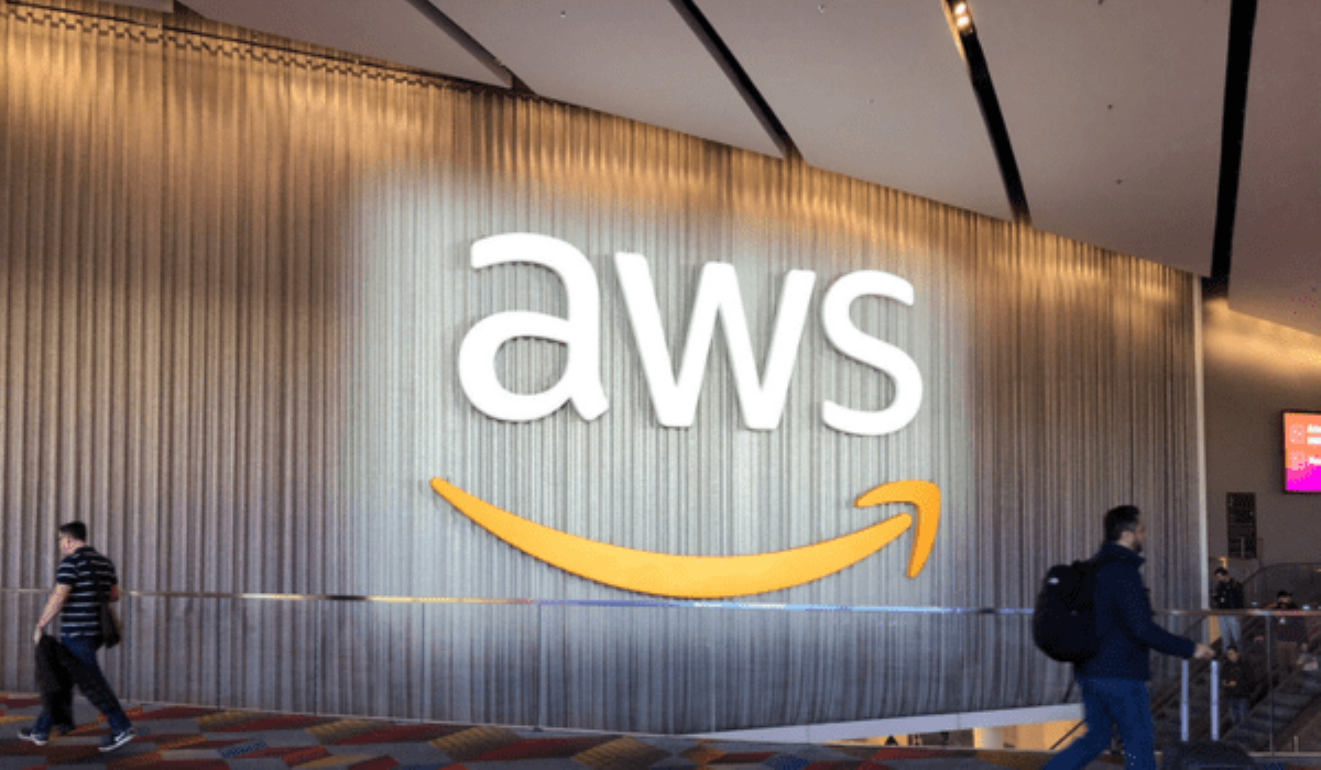 Large illuminated AWS logo displayed on a curtain wall inside a modern conference venue, with people walking past.
