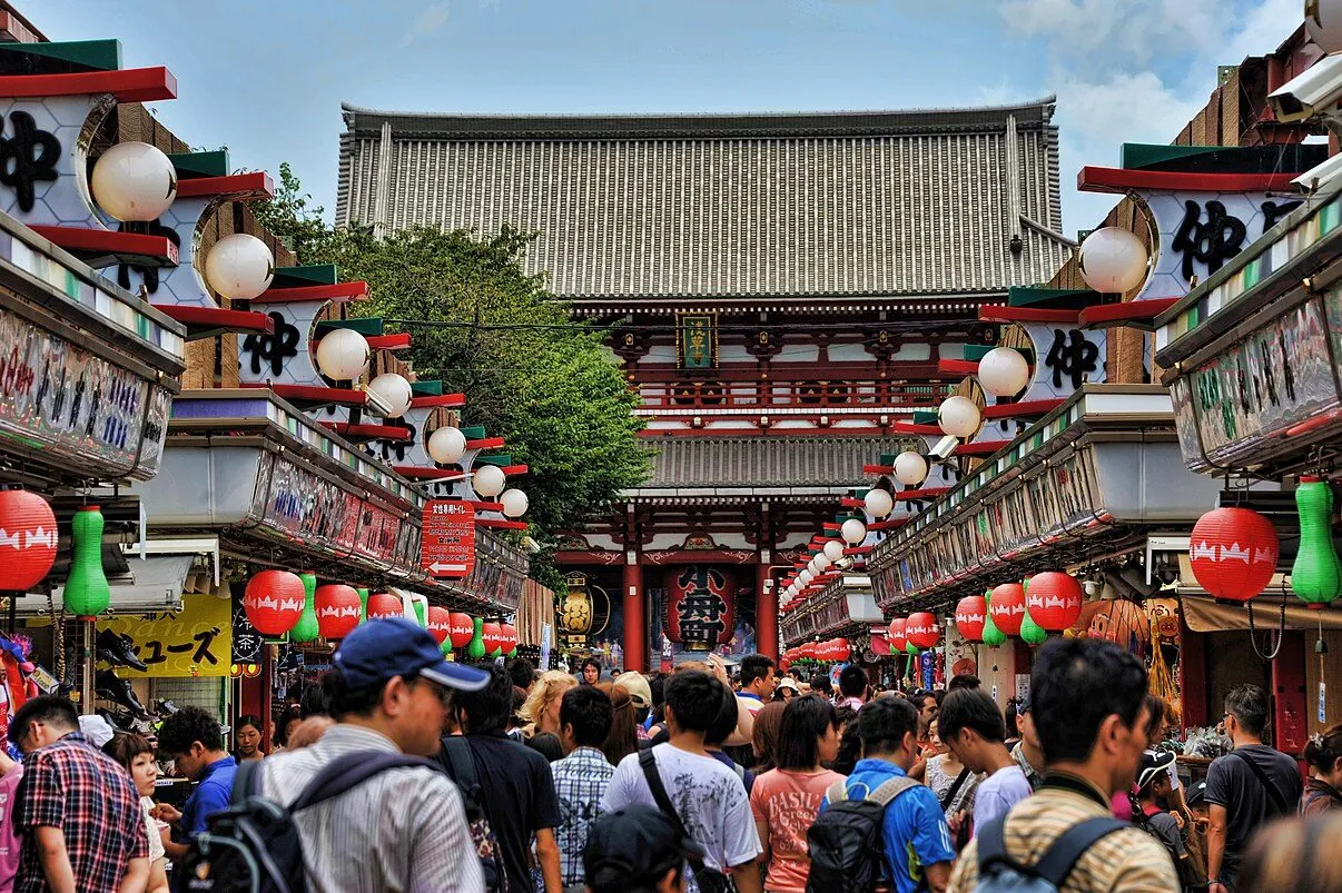 Crowds of tourists walk through Nakamise Shopping Street leading to Senso-ji Temple in Tokyo, Japan, surrounded by colorful lanterns and traditional signage.
