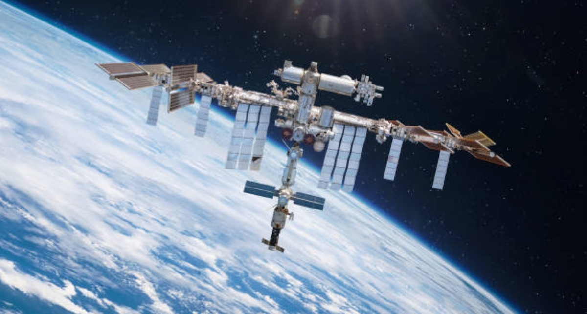 A high-resolution illustration of the International Space Station (ISS) set against the blackness of space. The ISS features multiple interconnected modules and large solar panel arrays extending symmetrically from both sides. Various components such as radiators, trusses, and docking ports are visible, showcasing the complexity and scale of the space station.