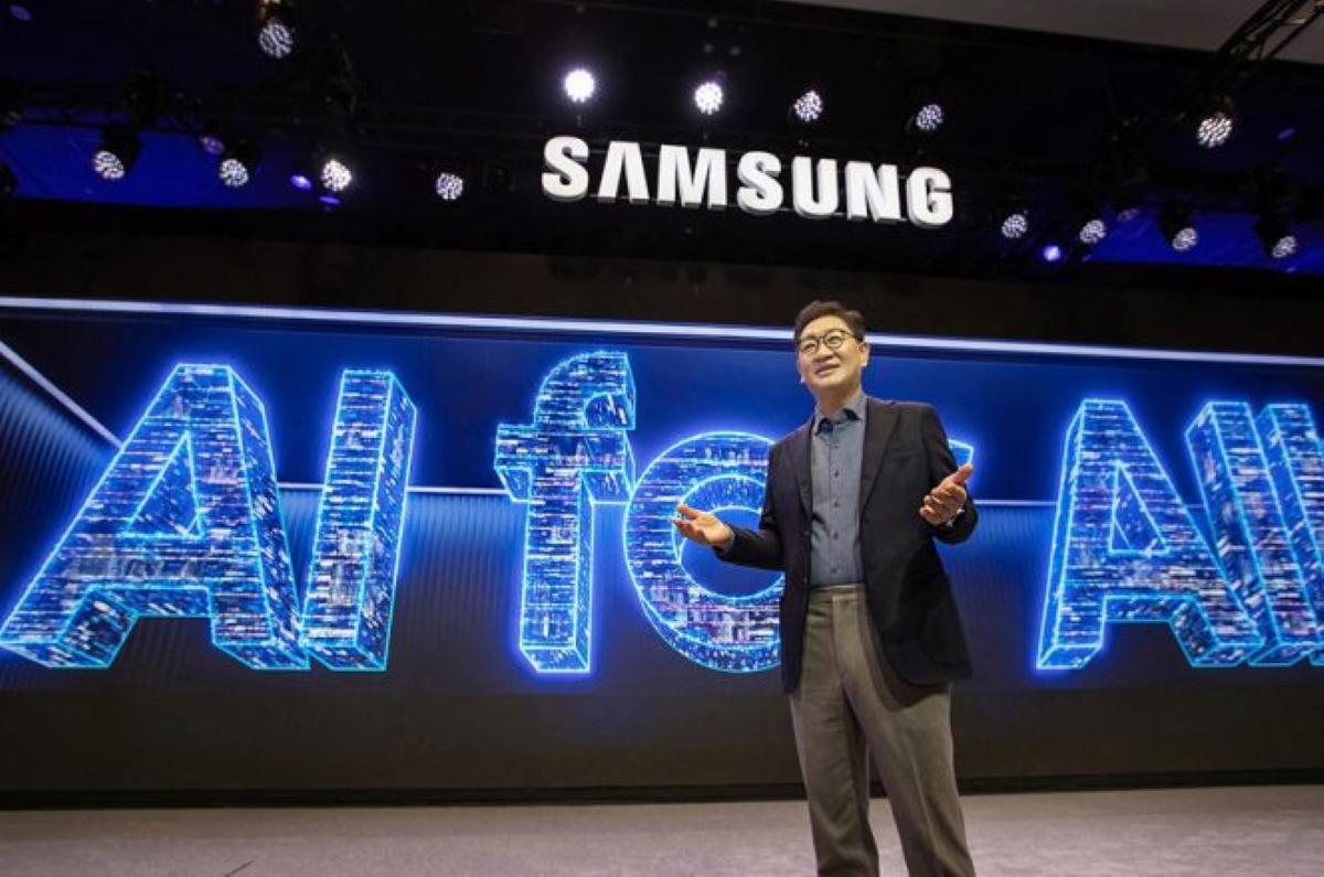A Samsung executive stands on stage delivering a keynote in front of a massive digital screen that reads "AI for All." The futuristic display features glowing blue letters with circuit-like patterns, emphasizing the tech company's focus on artificial intelligence accessibility. The stage is lit with overhead lights and the Samsung logo is prominently displayed above.