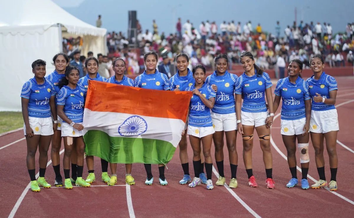 Indian women’s rugby sevens team celebrates on the track holding the national flag after a match, symbolizing teamwork, pride, and sporting achievement.