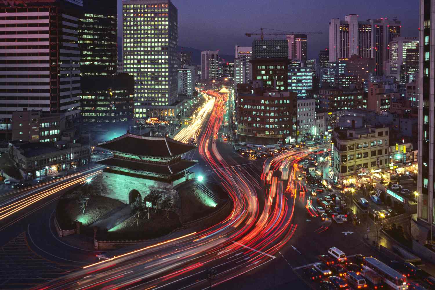 Night view of downtown Seoul with illuminated skyscrapers, historic Namdaemun Gate, and vibrant light trails from busy city traffic, showcasing South Korea’s blend of tradition and modern urban growth.