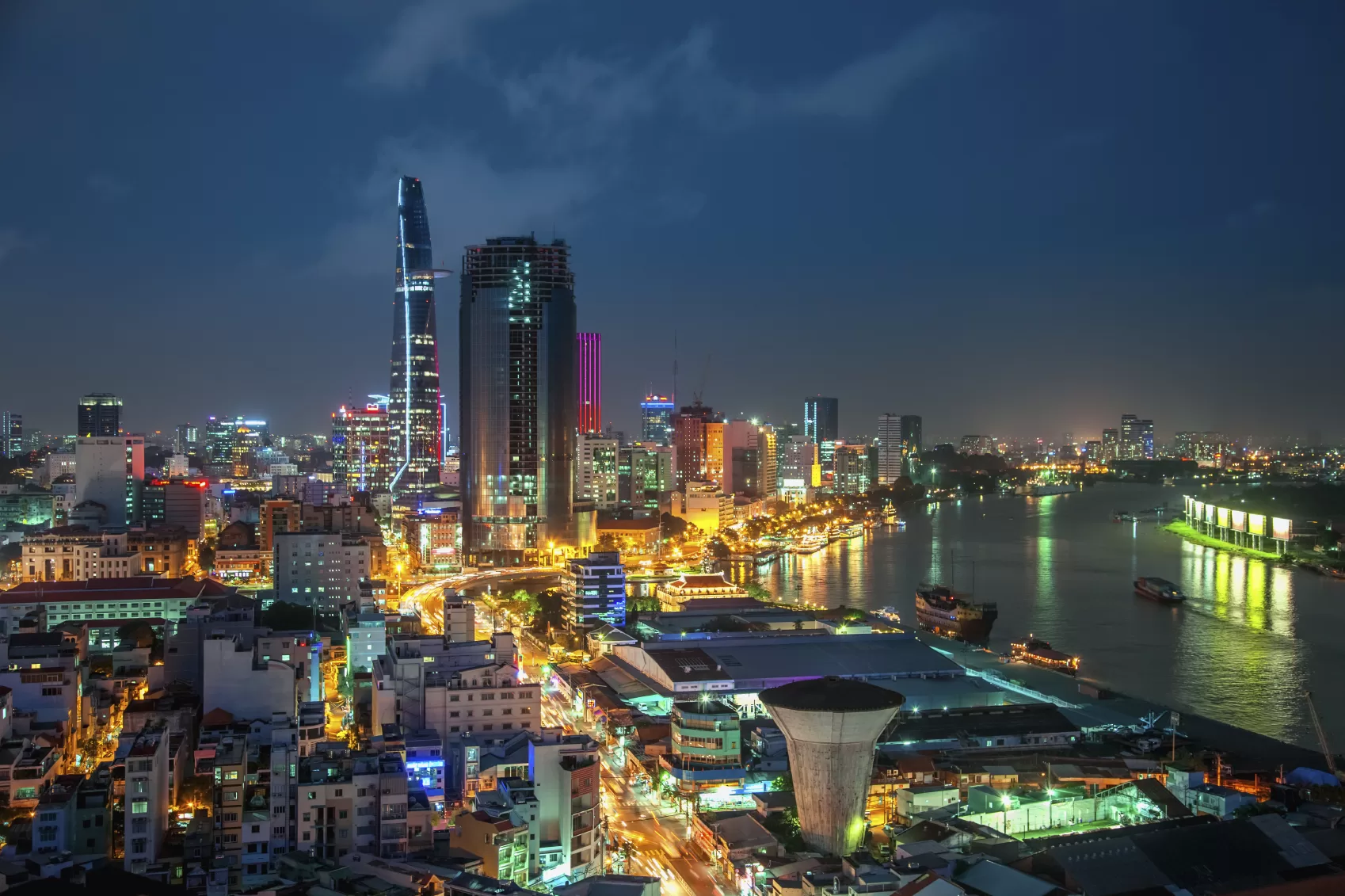 Night view of Ho Chi Minh City skyline with illuminated skyscrapers, including the Bitexco Financial Tower, alongside the Saigon River, showcasing vibrant city lights and reflections on the water.