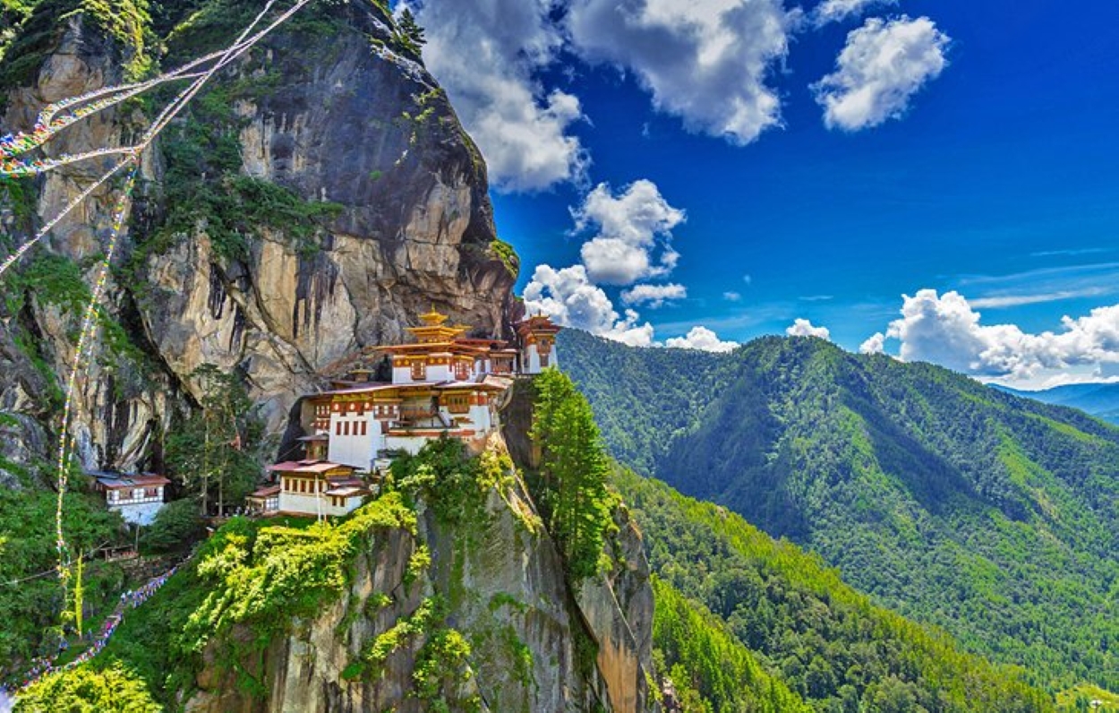 Paro Taktsang, also known as Tiger's Nest Monastery, perched dramatically on a cliffside in the Paro Valley, Bhutan, surrounded by lush green mountains under a bright blue sky with scattered clouds.