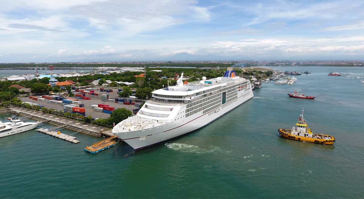 Aerial view of a luxury cruise ship docked at Benoa Harbour in Bali, Indonesia, with a yellow tugboat assisting in clear turquoise waters, surrounded by shipping containers and coastal infrastructure under a bright, partly cloudy sky.