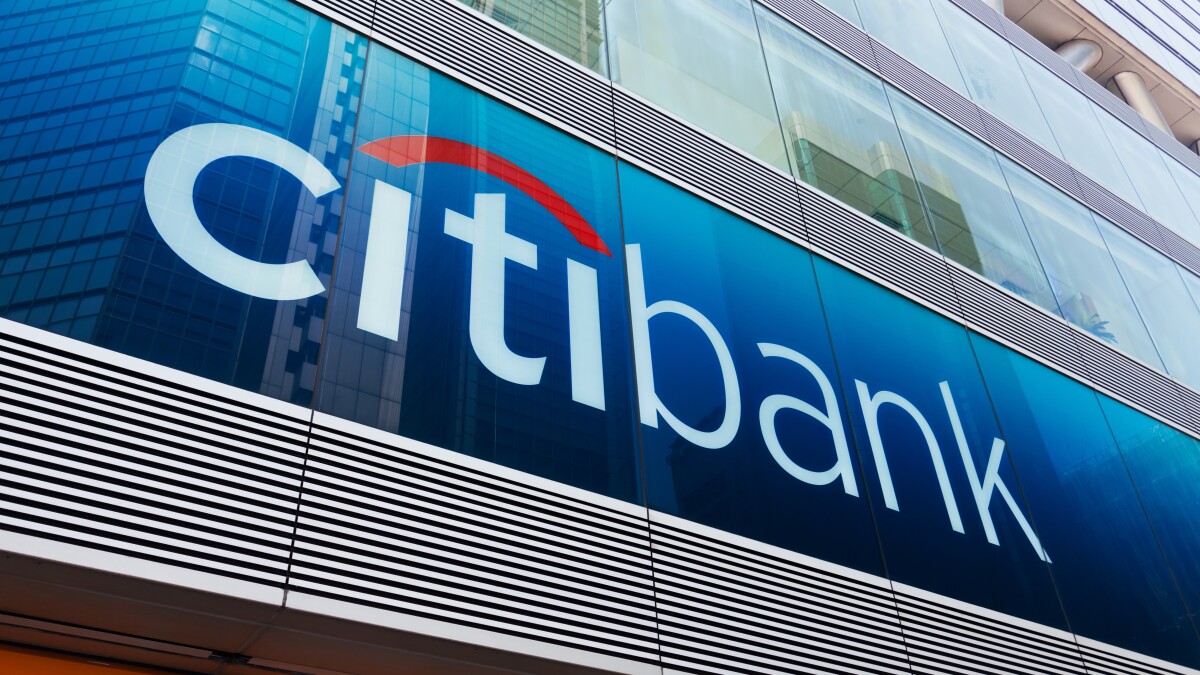 Citibank logo on the exterior glass facade of a modern urban office building, reflecting surrounding skyscrapers in a financial district.