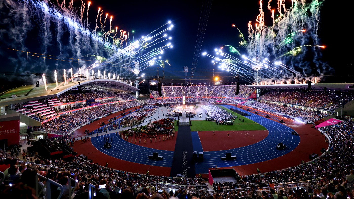 Fireworks light up the night sky at a packed stadium during a grand sports event opening ceremony, with thousands of spectators in attendance and vibrant colors creating a festive atmosphere.
