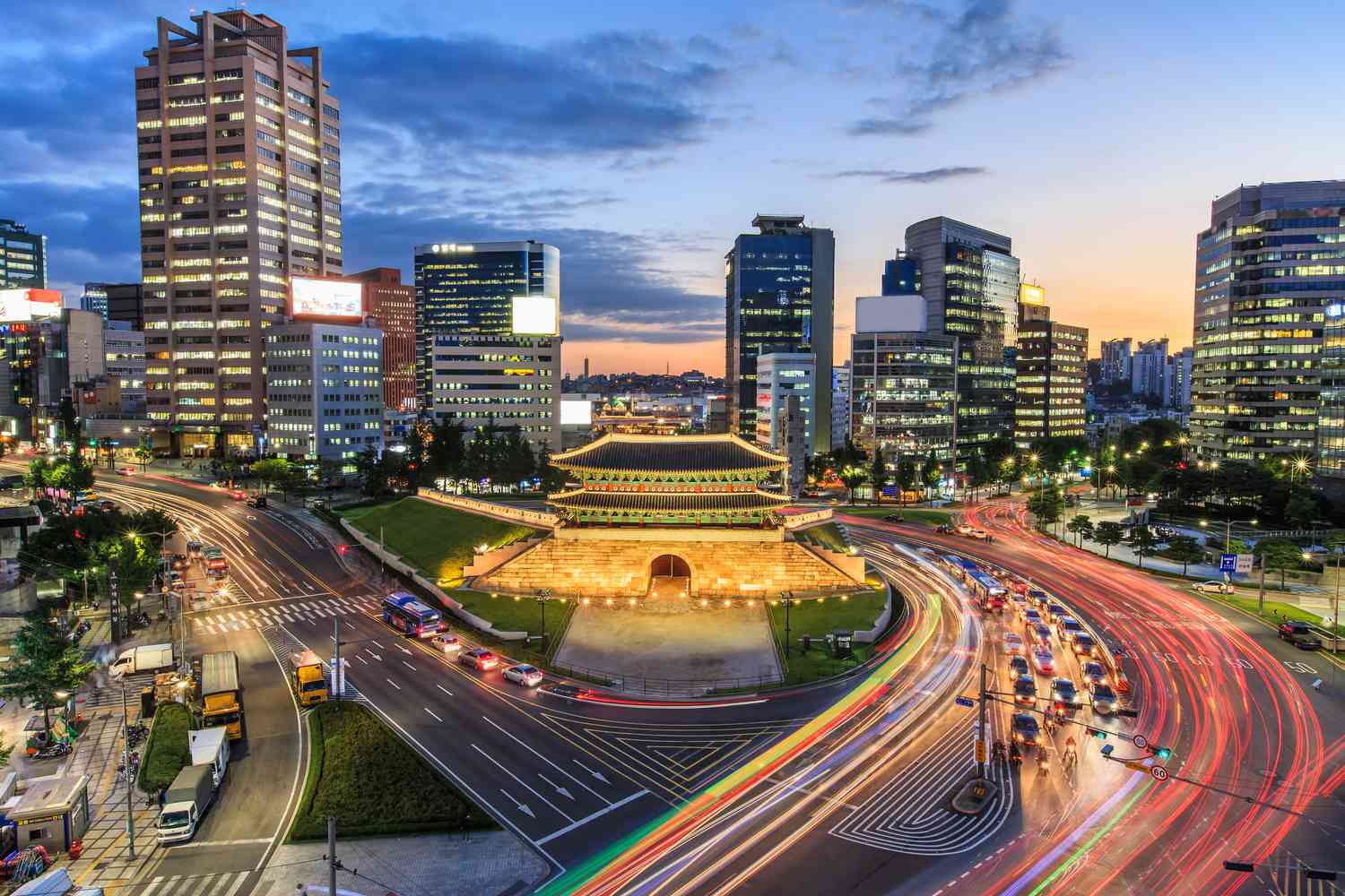 Night view of Seoul’s Namdaemun Gate surrounded by illuminated modern skyscrapers and busy traffic trails, showcasing South Korea’s blend of history and urban growth.