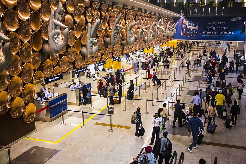 Travelers queue at immigration counters inside Indira Gandhi International Airport in Delhi, featuring a decorated wall with giant mudra hand sculptures and golden discs, symbolizing India’s cultural heritage in a modern airport setting.