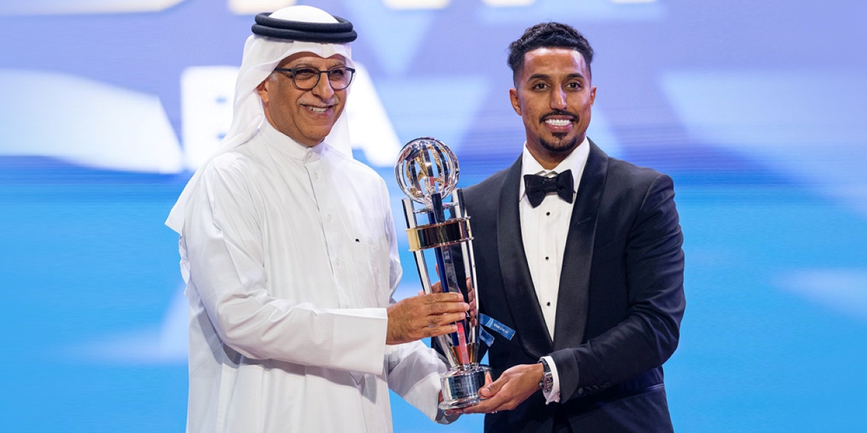 A football player in a tuxedo receives a major sports award trophy from an official in traditional Gulf attire during a ceremony on stage.