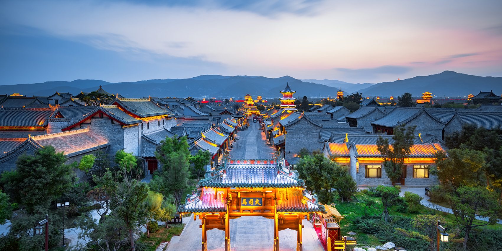 Evening view of Pingyao Ancient City in Shanxi, China, with traditional Chinese architecture illuminated under twilight and surrounded by distant mountains.