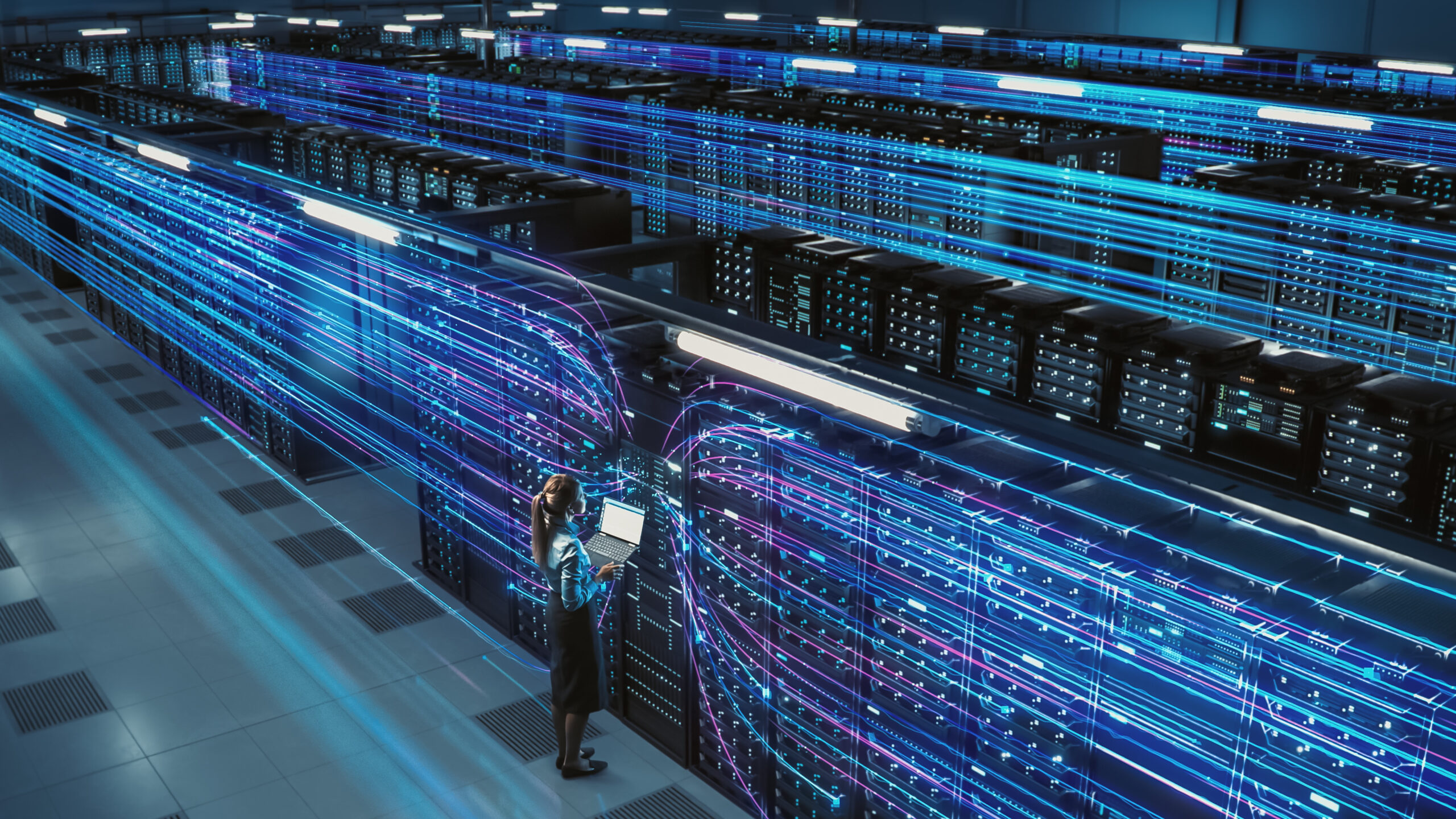 Female engineer working with laptop in a futuristic data centre, surrounded by illuminated servers and digital network lines representing high-speed data transfer and cloud computing.