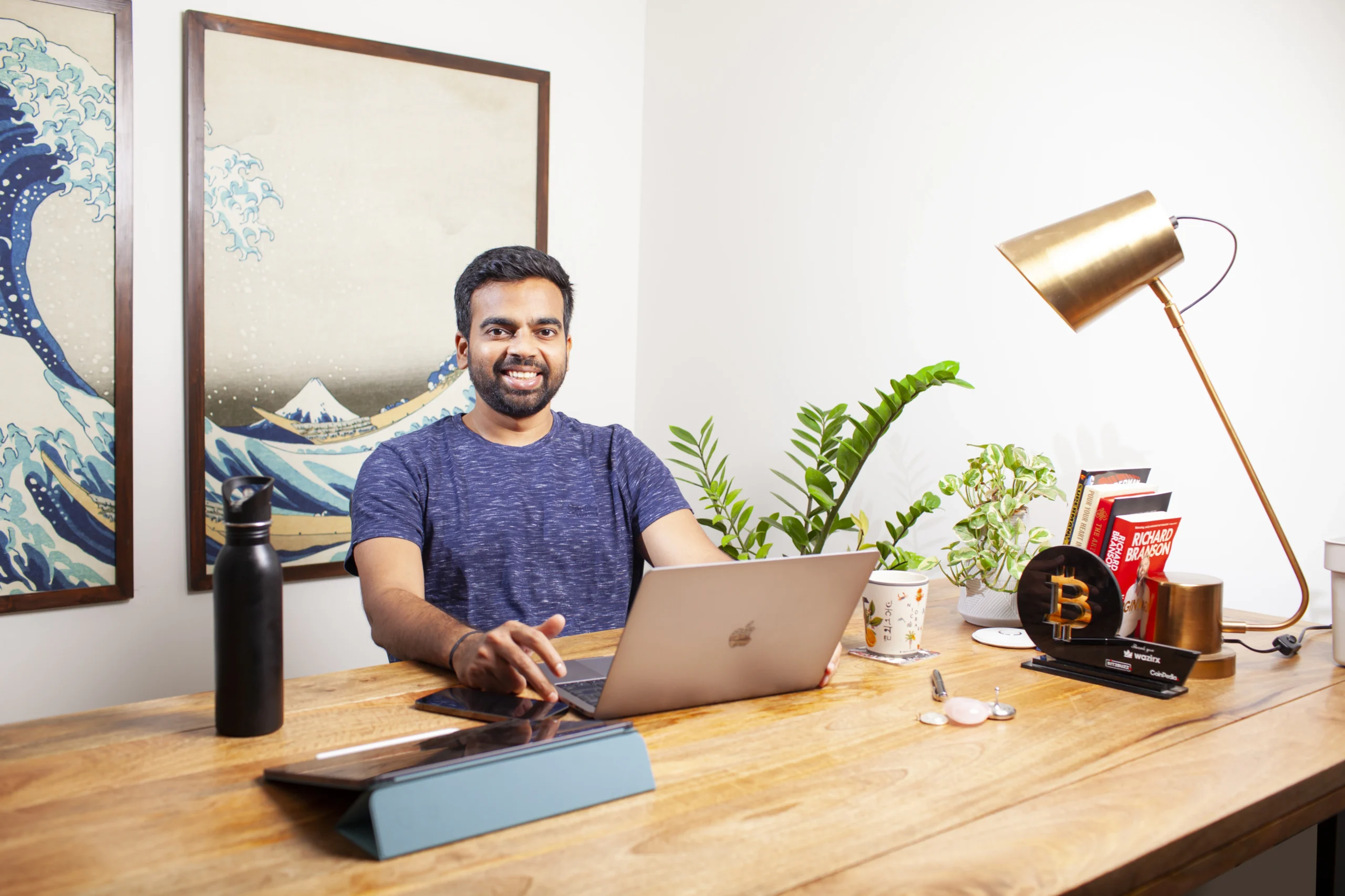 A man working at a modern wooden desk with a laptop, surrounded by books, plants, and decor, creating a professional home office setup with a creative touch.