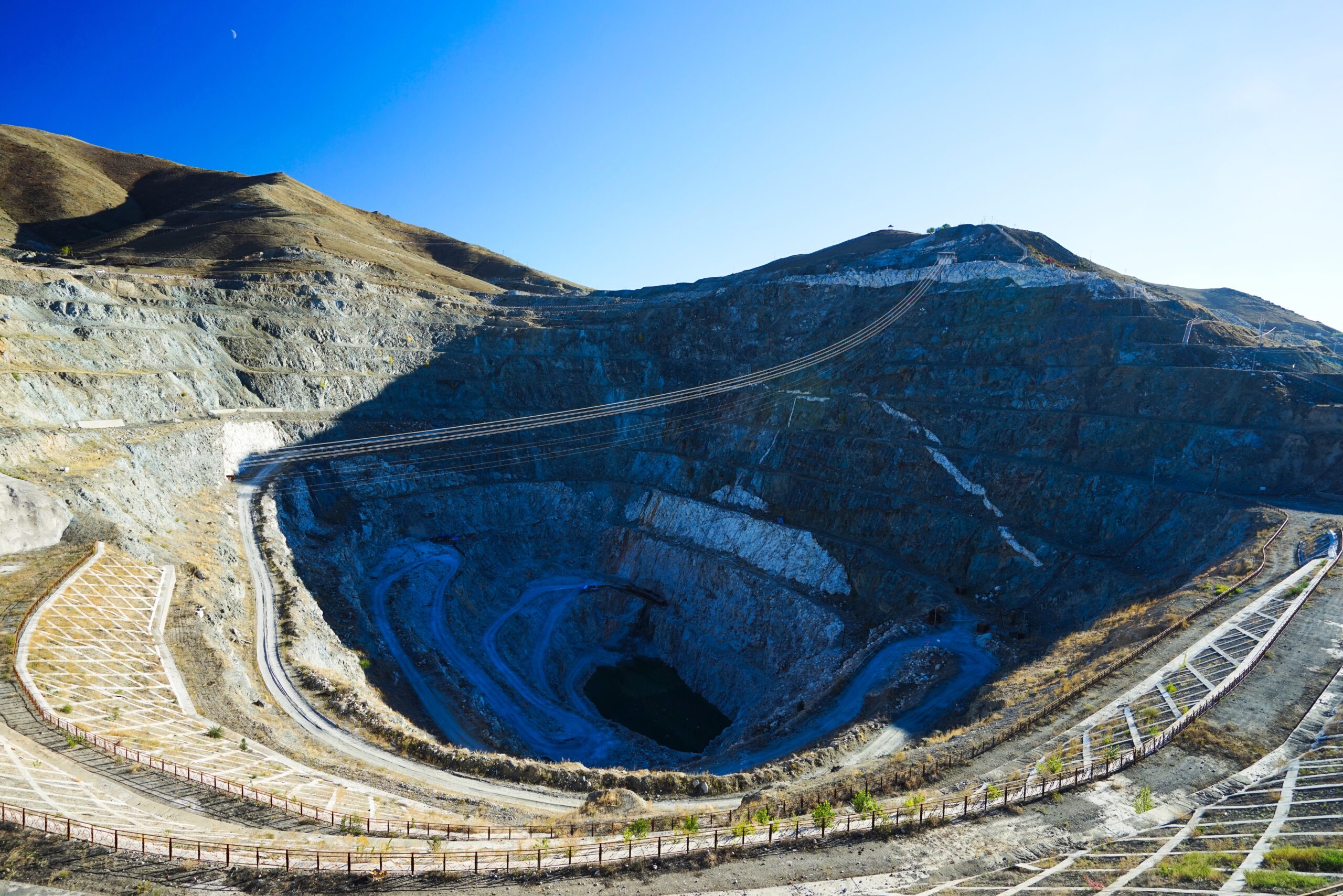 Open-pit mining site surrounded by rocky terrain and hills under a clear blue sky, showcasing large-scale mineral extraction operations.