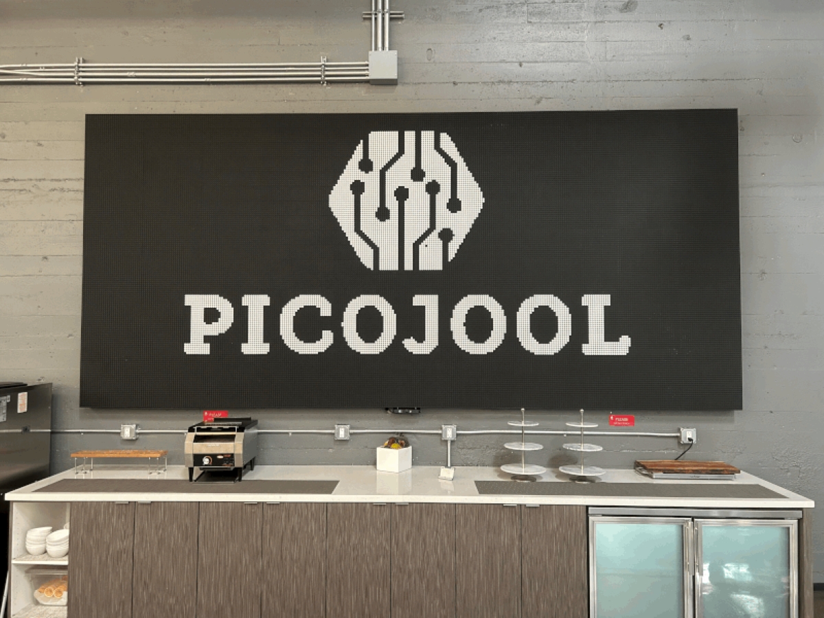 Modern office kitchenette with a large black wall display showing the PICOJOOL logo featuring a circuit-inspired icon above the brand name.