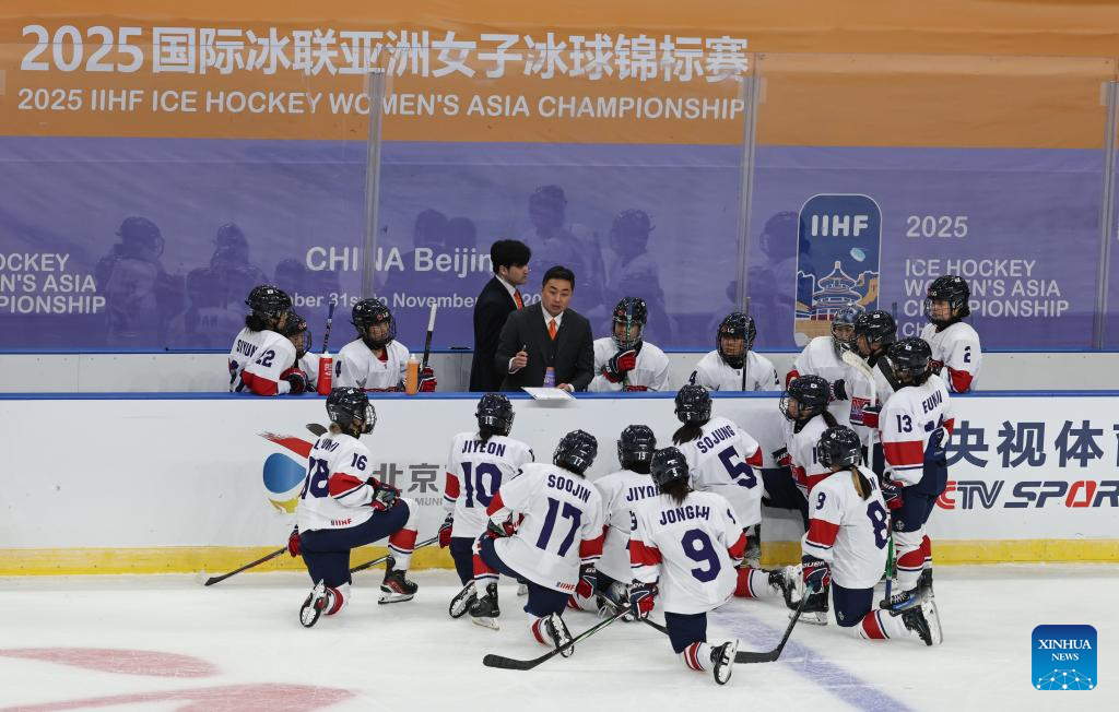 Women’s ice hockey team gathered around their coach during a timeout at the 2025 IIHF Ice Hockey Women’s Asia Championship in Beijing, with players in white and navy uniforms listening closely at the rinkside bench.