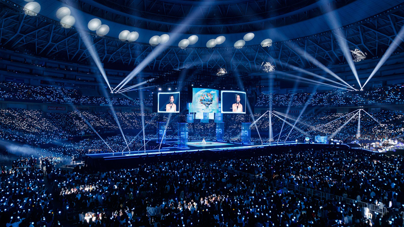 Packed indoor stadium lit with blue light sticks during a large-scale concert, featuring a central stage, towering screens, and dramatic spotlights illuminating the performer.