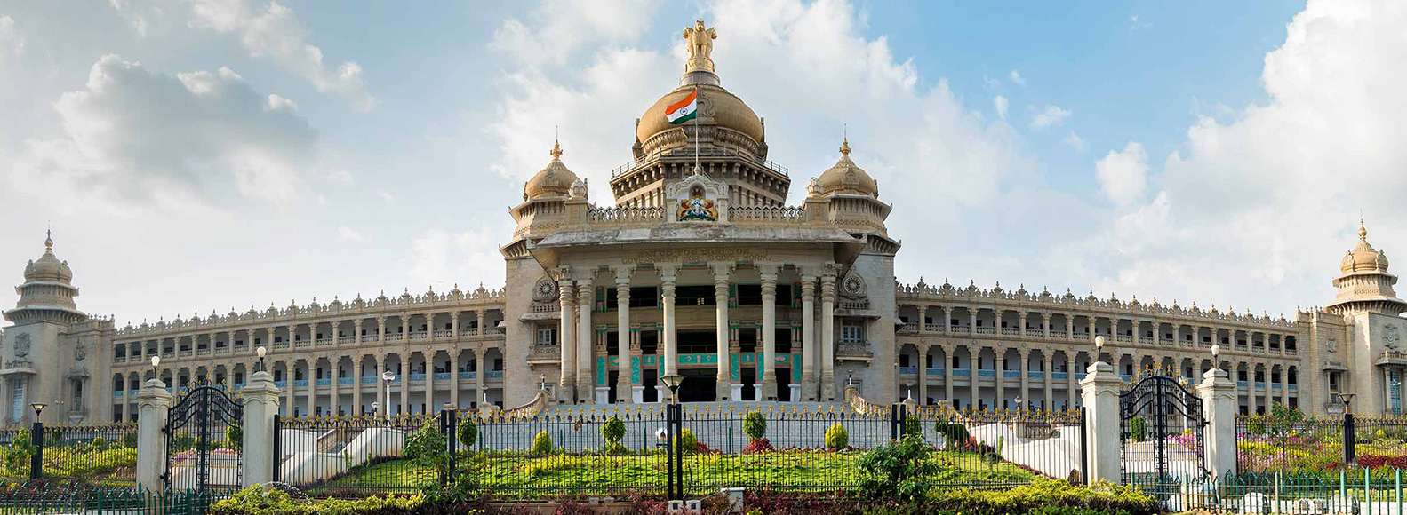 Panoramic view of Vidhana Soudha in Bengaluru, India, showcasing its grand Neo-Dravidian architecture, central dome with the Indian flag, and expansive landscaped grounds under a bright sky.