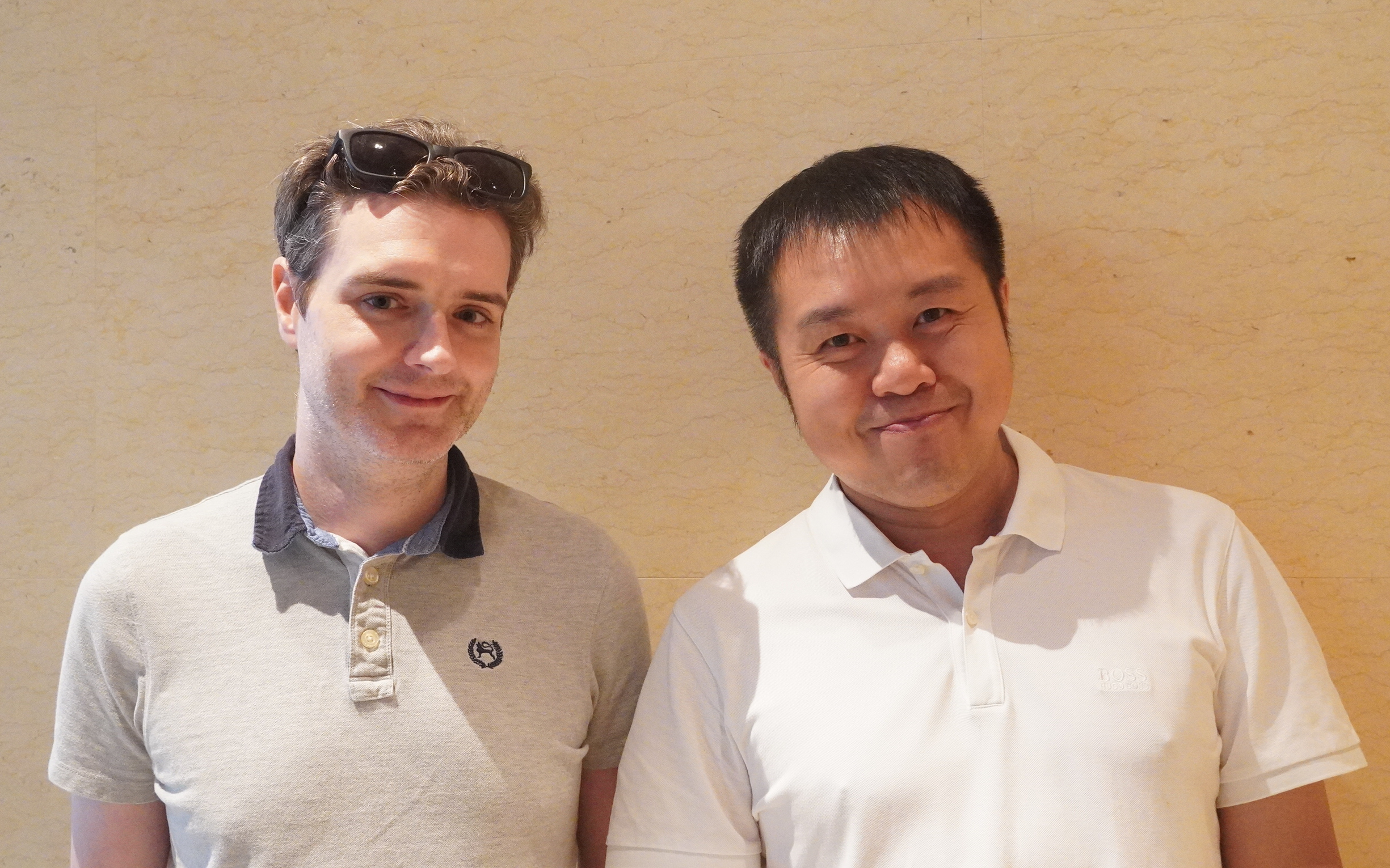 Two men standing side by side against a beige wall, smiling casually while wearing light-colored polo shirts.