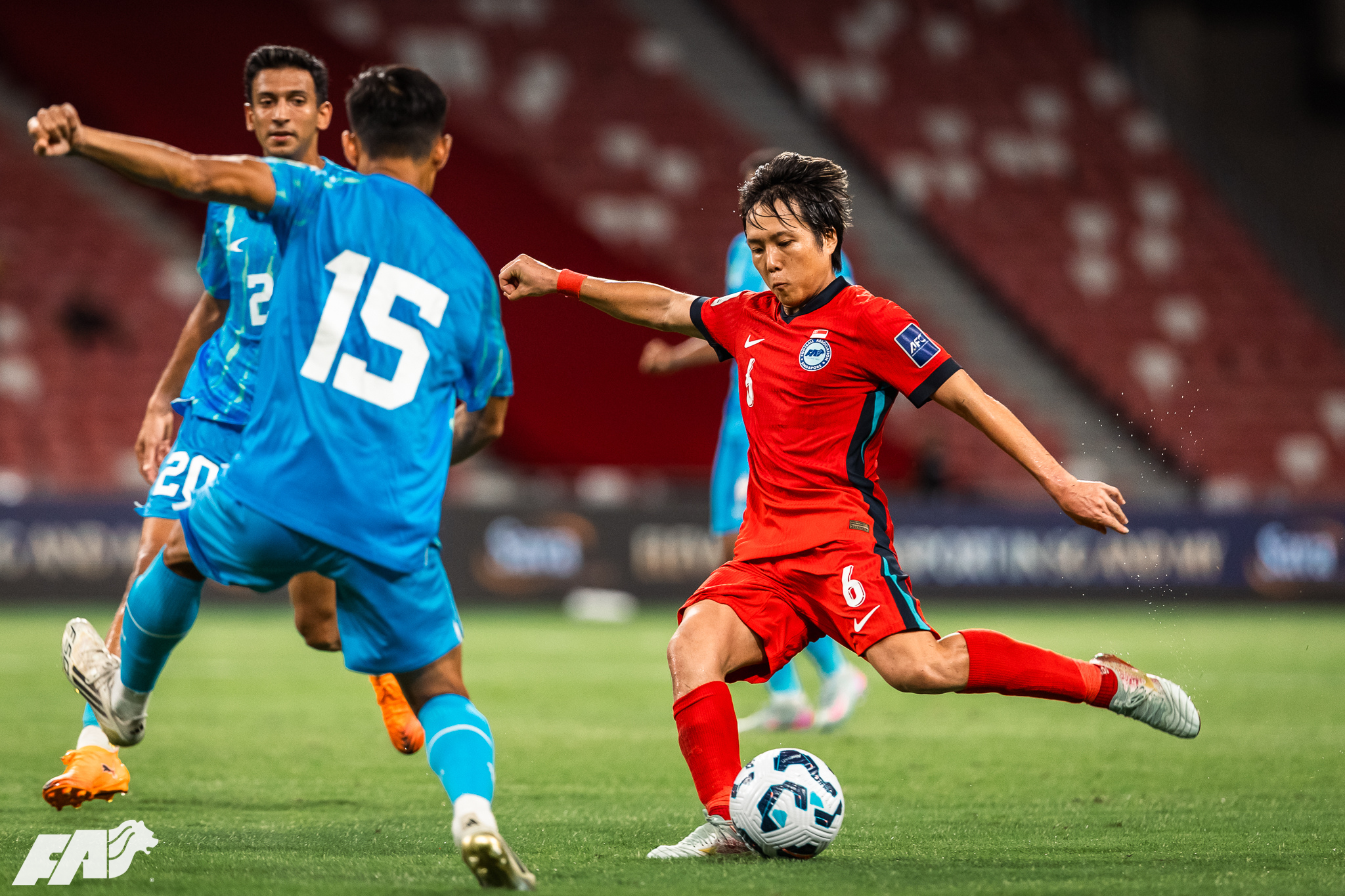 Women’s international football match action, with a player in a red jersey striking the ball while two opponents in blue kits close in during a competitive midfield challenge.