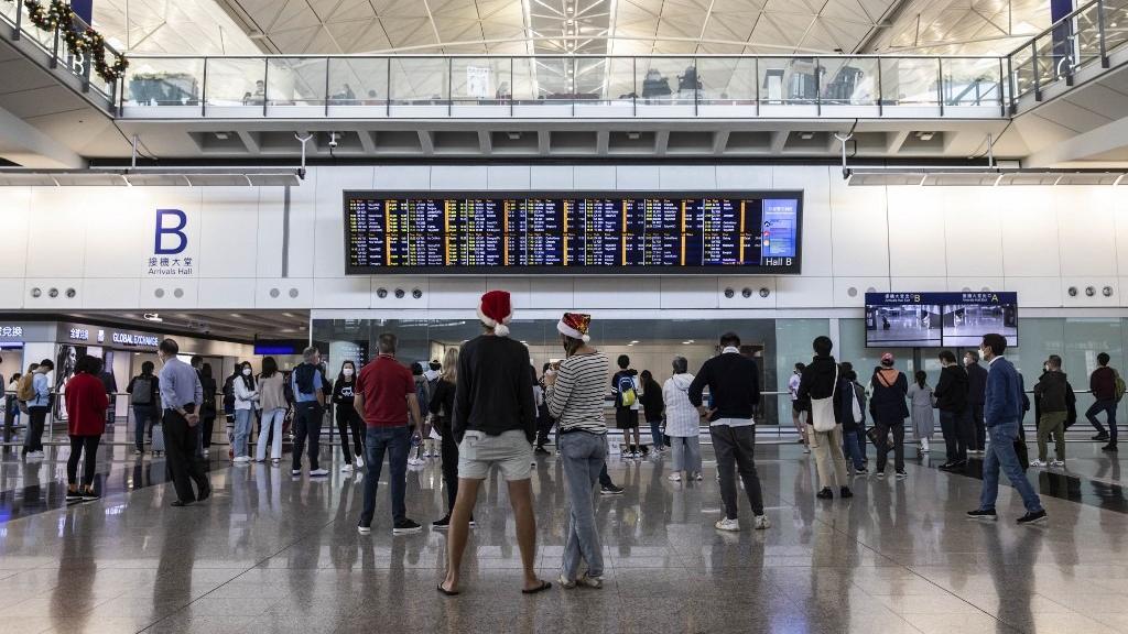 Passengers gathered inside an international airport arrivals hall, checking flight information boards amid busy travel activity.