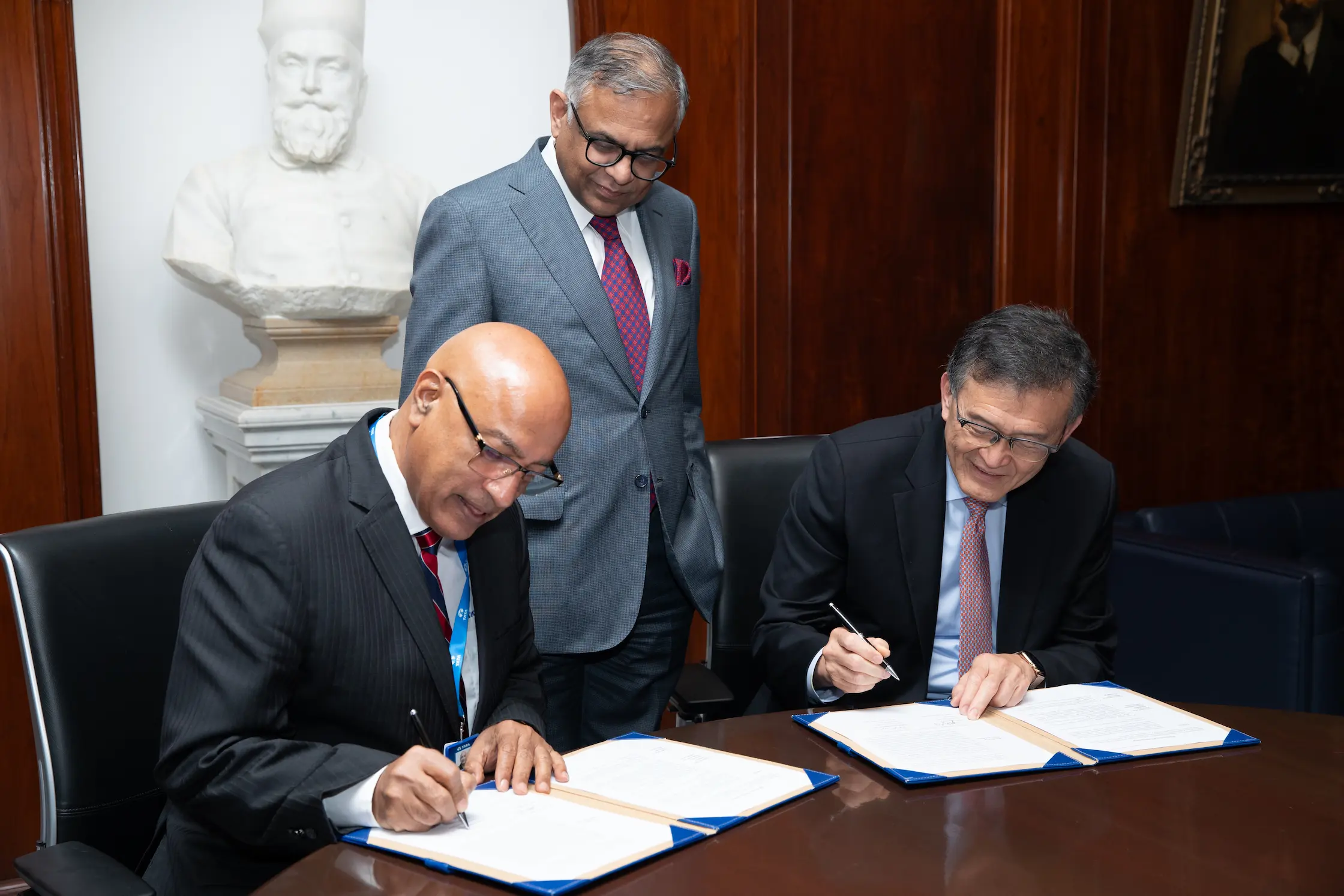 Two executives in dark suits sign documents at a boardroom table while a third senior official in a grey suit looks on, marking a formal partnership agreement.