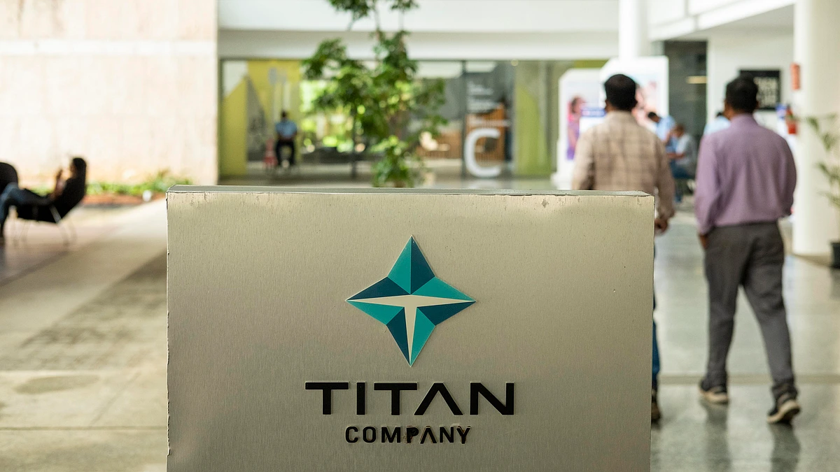 Titan Company logo displayed on a brushed metal sign inside a modern corporate campus, with employees walking through a bright, open office courtyard in the background.