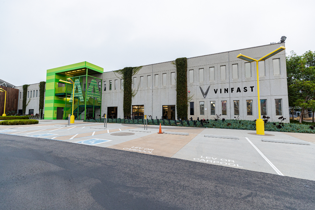 Exterior view of a VinFast corporate facility featuring a modern concrete facade, green-accented entrance structure, VinFast logo signage, and designated electric vehicle carpool parking in front.