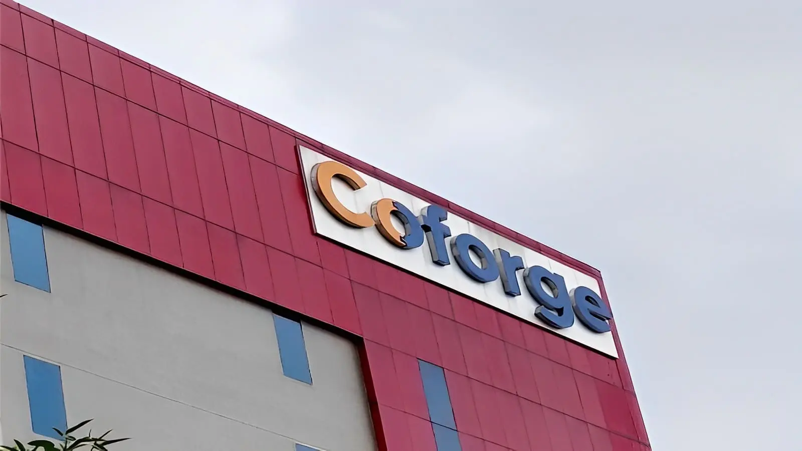 Coforge corporate logo mounted on the exterior of a modern office building with a red and grey facade under an overcast sky.