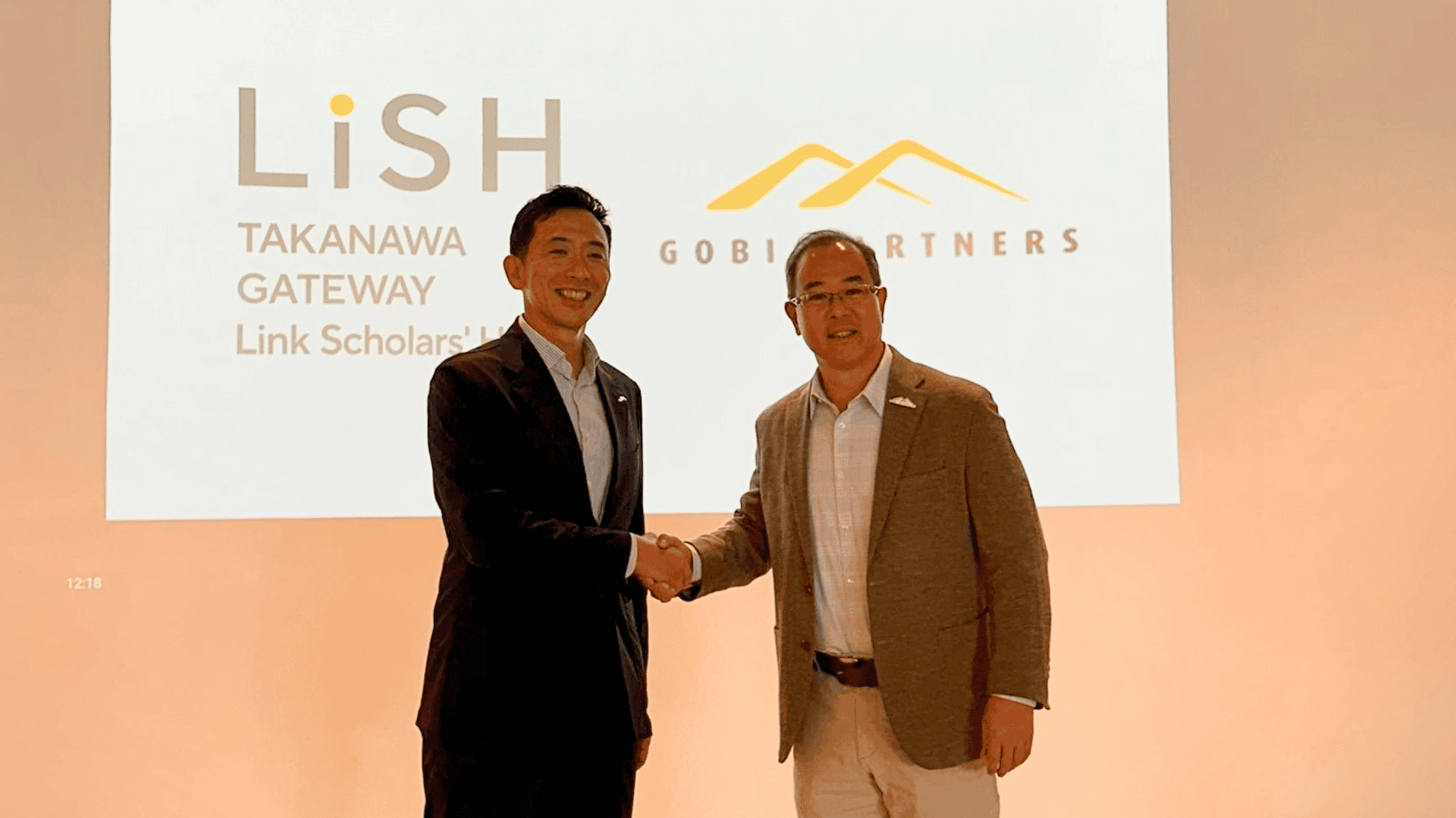 Two business leaders shaking hands on stage during a partnership announcement, with LiSH Takanawa Gateway and Gobi Partners logos displayed on a presentation screen in the background.