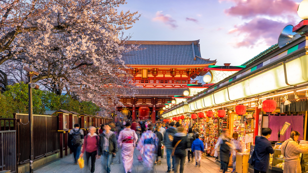 Crowded market street in Asakusa, Tokyo, lined with lantern-lit stalls and cherry blossoms, with Sensō-ji Temple gate in the background at sunset.