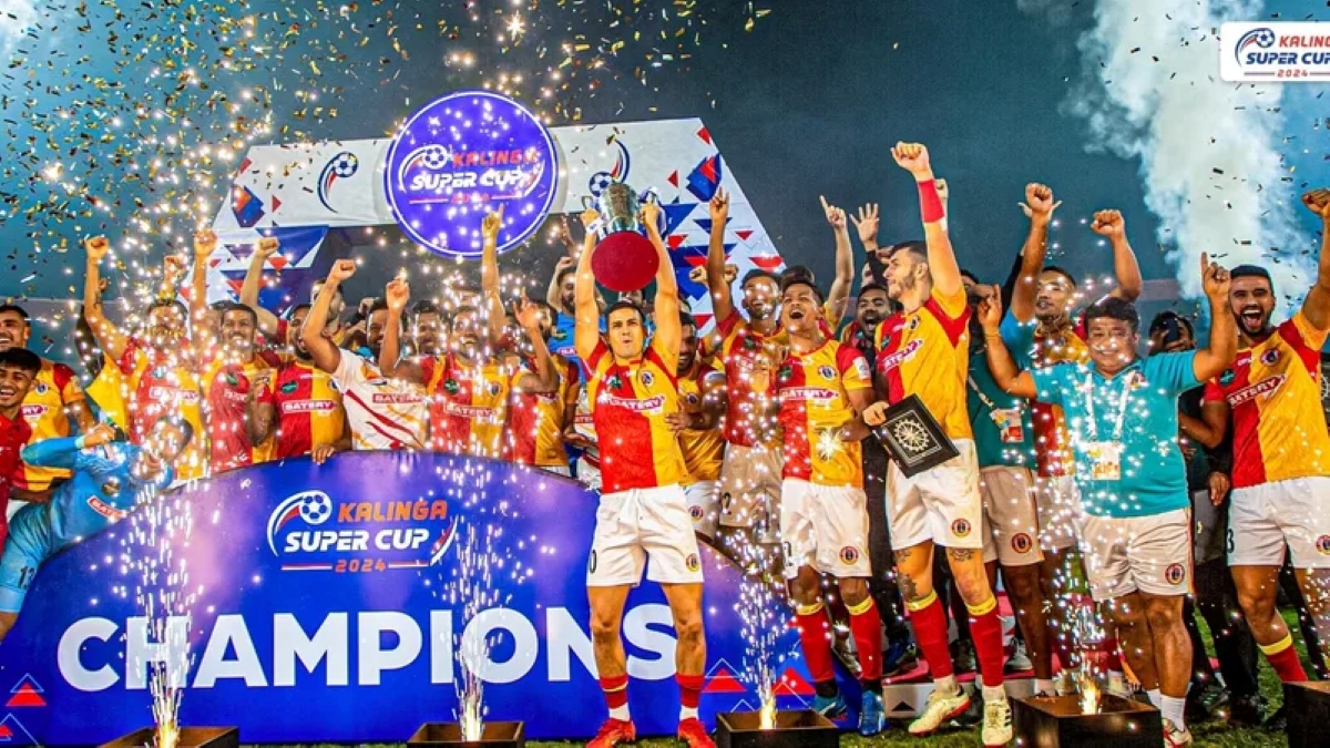 Football team celebrating Kalinga Super Cup 2024 title, lifting trophy on “Champions” stage with confetti and fireworks.