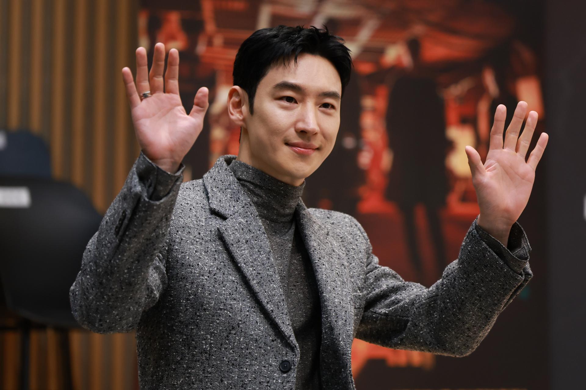 Man in a grey blazer and turtleneck smiling and raising both hands in greeting at an indoor event, with a softly blurred stage or poster backdrop behind him.