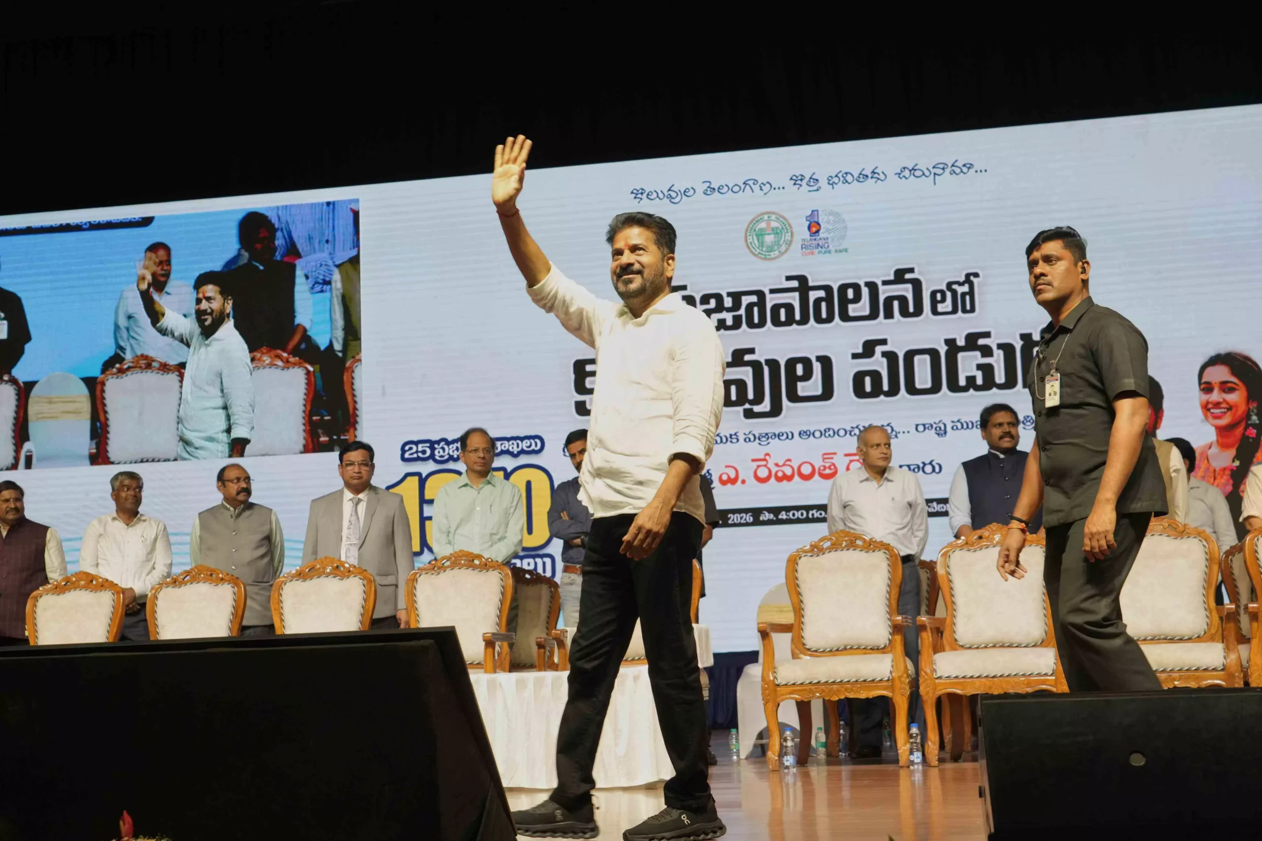 Indian political leader waving to the audience on stage during a public event, with officials seated behind and a large backdrop highlighting a government or civic programme, reflecting public engagement and regional leadership in India.