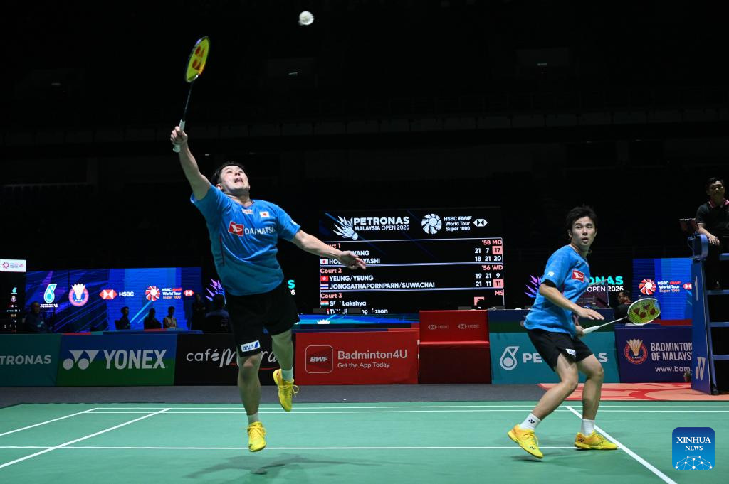 Men’s doubles badminton match in action at an international BWF tournament, with two players in blue jerseys executing an overhead smash and defensive return on an indoor court during a high-intensity rally.