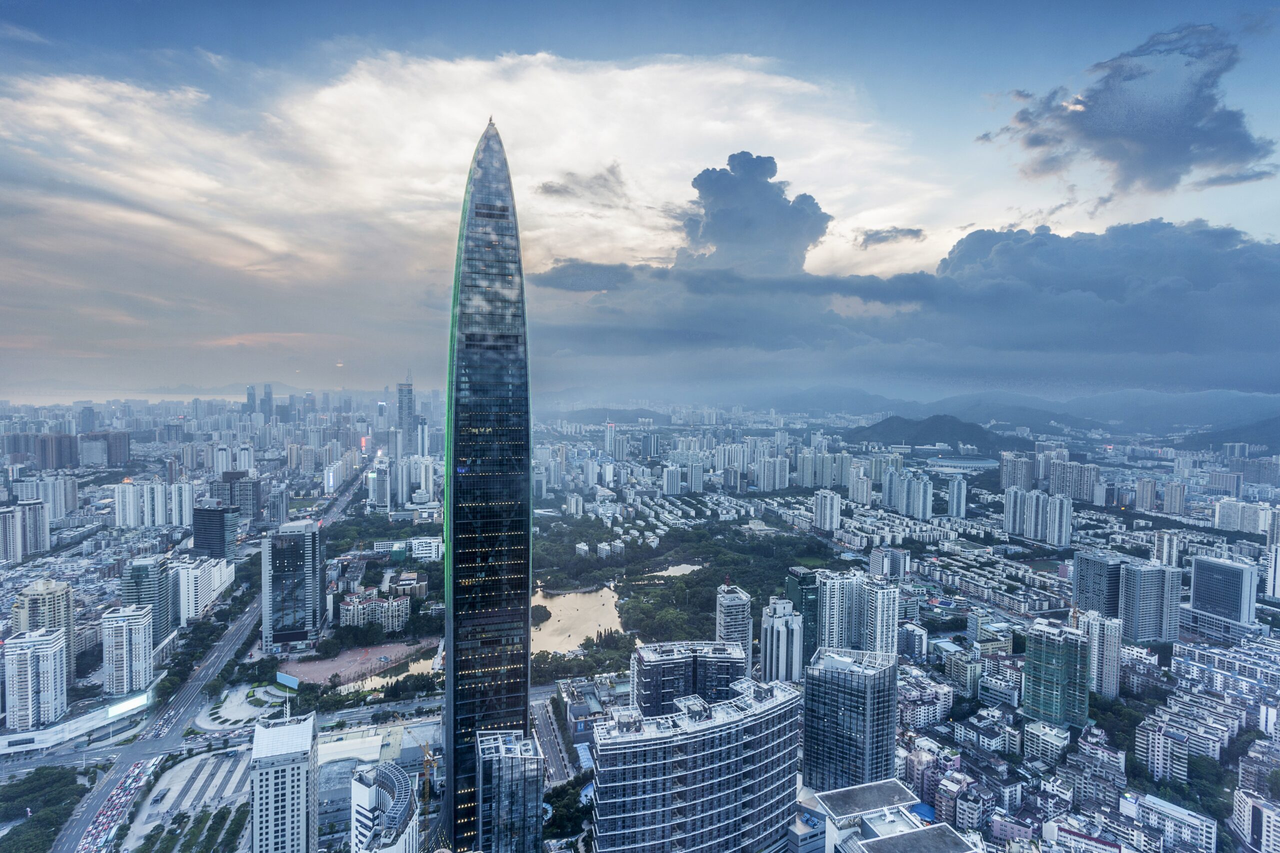 Aerial view of Shenzhen’s city skyline at dusk, featuring the Ping An Finance Center rising above dense urban districts, modern high-rise buildings, and surrounding hills under dramatic cloud formations.