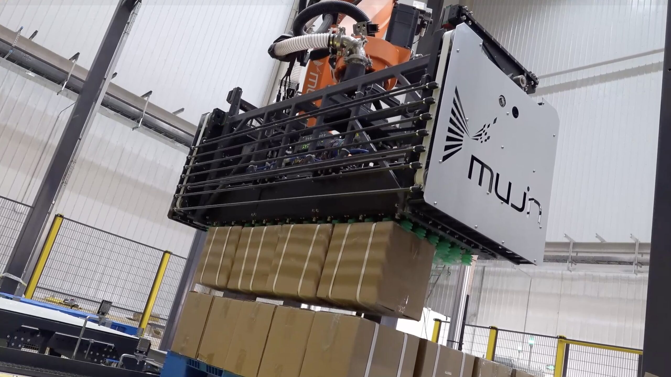 Industrial robotic palletizing system lifting a stack of cardboard boxes, featuring a Muju-branded robotic gripper operating inside a warehouse automation facility.