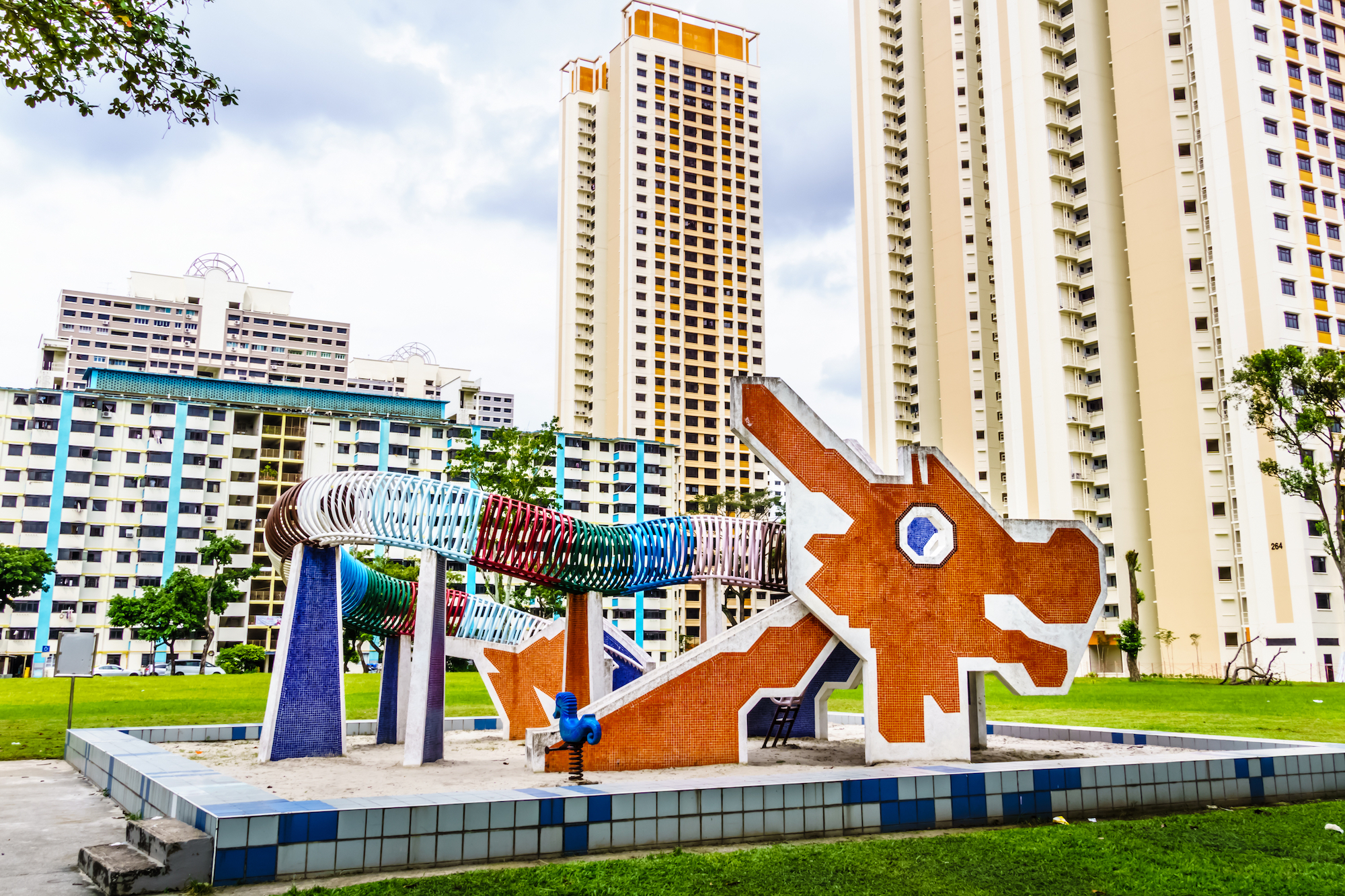 Dragon-themed playground sculpture set within a public housing estate, surrounded by high-rise residential apartment blocks and green open space, reflecting urban community design and family-friendly living.