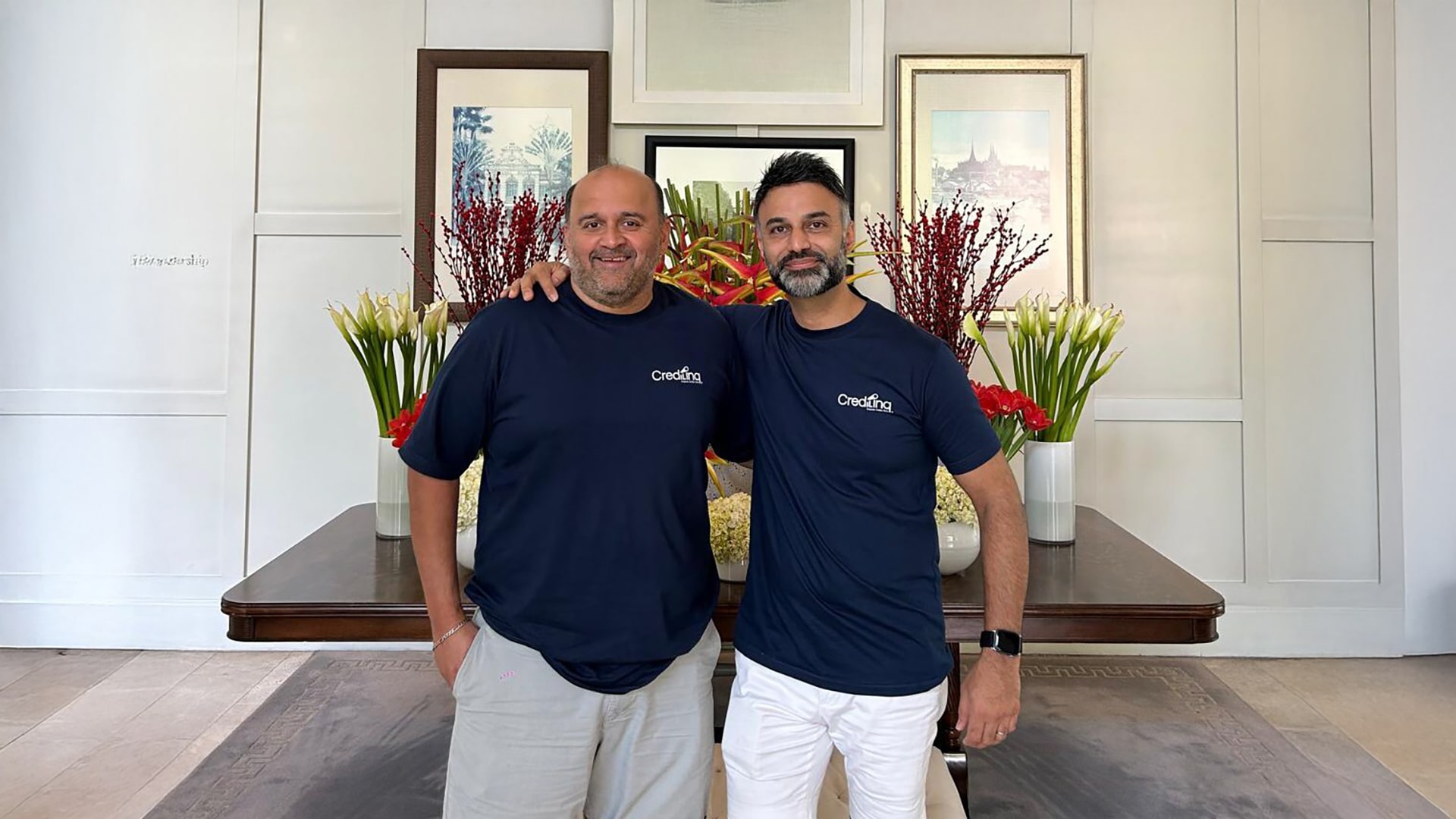 Two startup founders standing side by side indoors, wearing branded company T-shirts, posing in front of a floral arrangement and framed artwork, symbolis