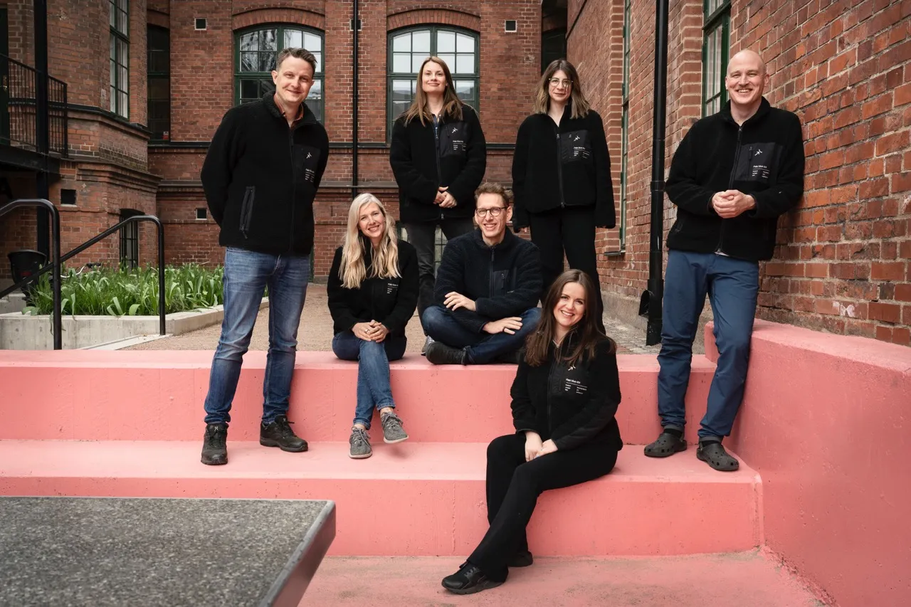 Startup team group portrait outdoors, with founders and staff wearing black jackets, seated and standing on pink steps in a brick courtyard, representing modern workplace culture, collaboration, and tech innovation.