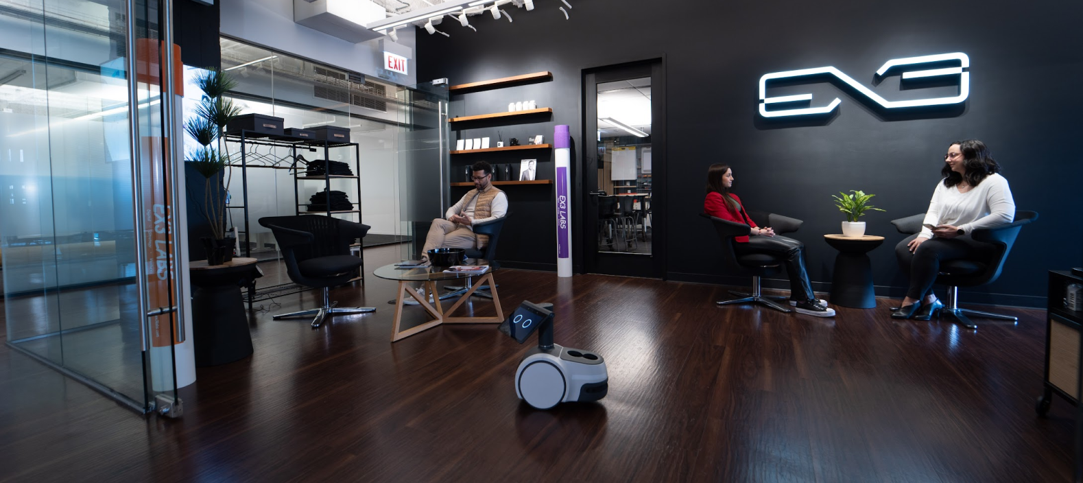 Modern startup office lounge featuring a small autonomous service robot navigating a meeting area, with professionals seated in discussion under a minimalist tech logo, highlighting workplace robotics, human–AI interaction, and next-generation office automation.