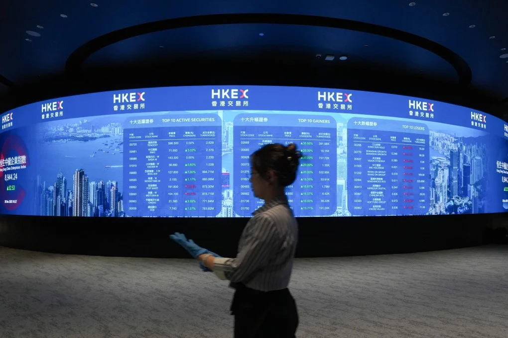 Hong Kong Exchange (HKEX) digital stock board displaying top gainers and active securities, with a staff member walking past in a modern financial trading hall.