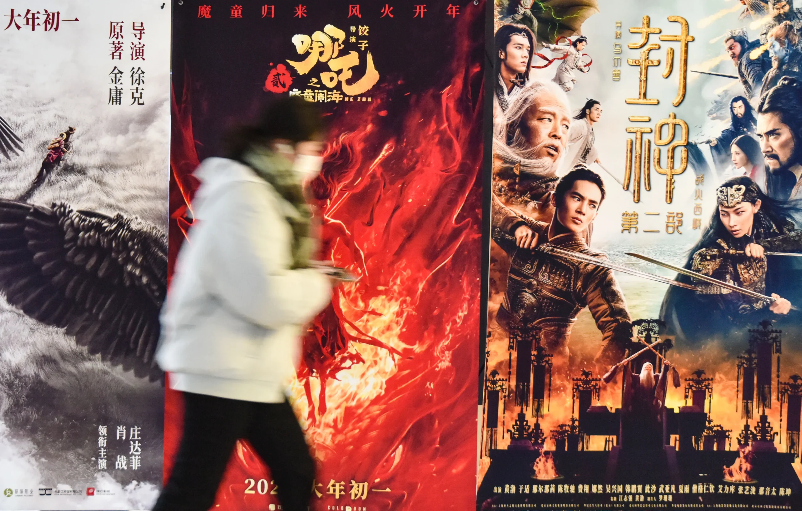 Pedestrian walks past colorful Chinese movie posters promoting fantasy and historical blockbuster films in an urban cinema display.