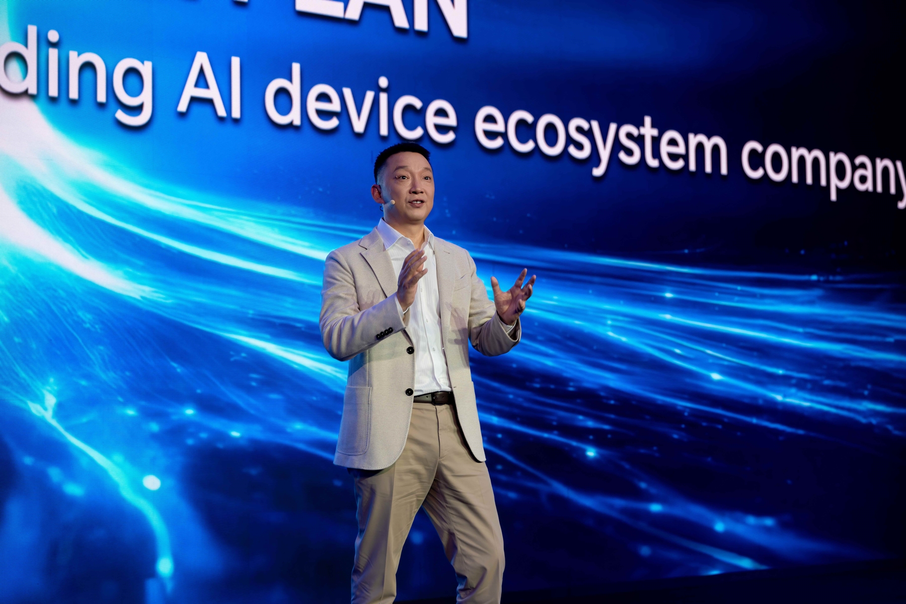 Technology executive delivering keynote on building AI device ecosystem at global innovation conference, speaking on stage with digital blue backdrop.