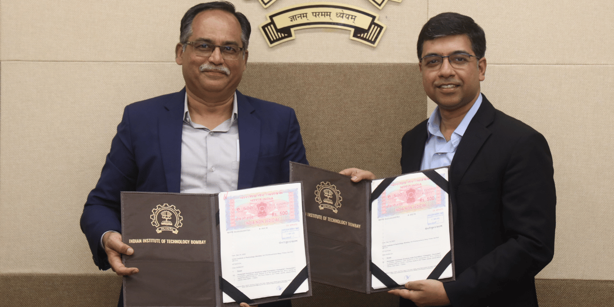 Two executives shaking hands while holding signed documents beneath an institutional emblem, symbolizing a partnership agreement between a company and a technology institute.