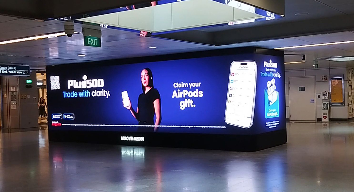 Large digital Plus500 trading platform advertisement displayed in a busy metro station, promoting mobile trading app and AirPods gift offer.